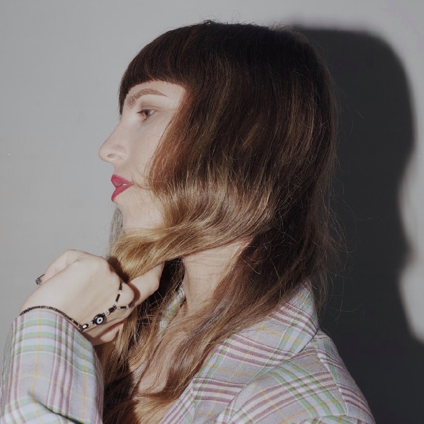 Sara is photographed from the side, looking away from the camera and wearing a plaid blazer.